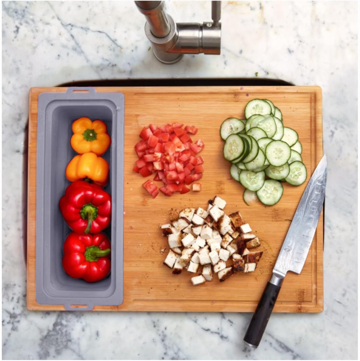 A bamboo cutting board with a knife on it, featuring prepped vegetables including sliced cucumbers, tomatoes, and bell peppers, and a blue removable strainer.